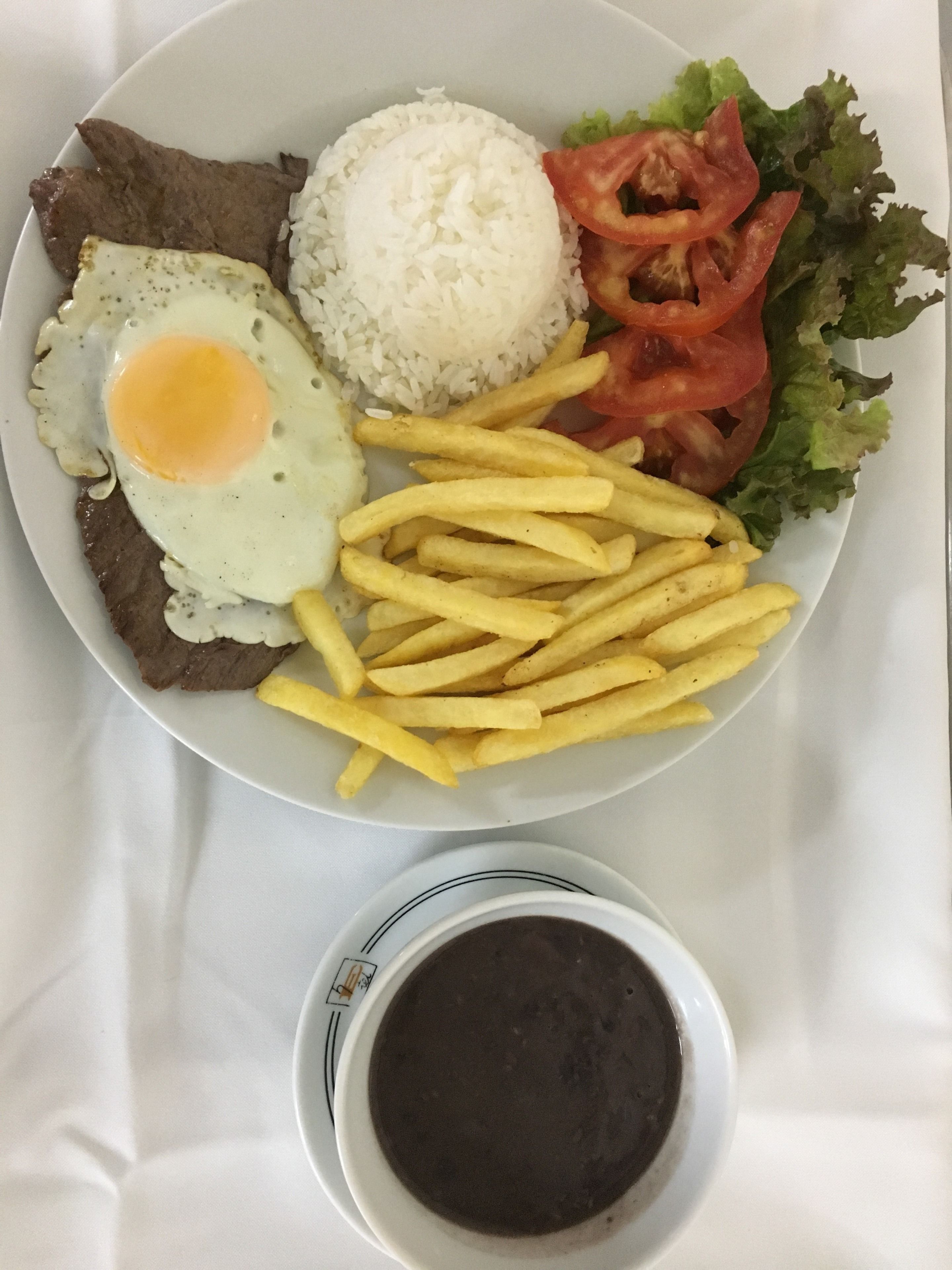 A plate with a fried egg, beef, rice, French fries, tomato slices, lettuce, and a small bowl of sauce.