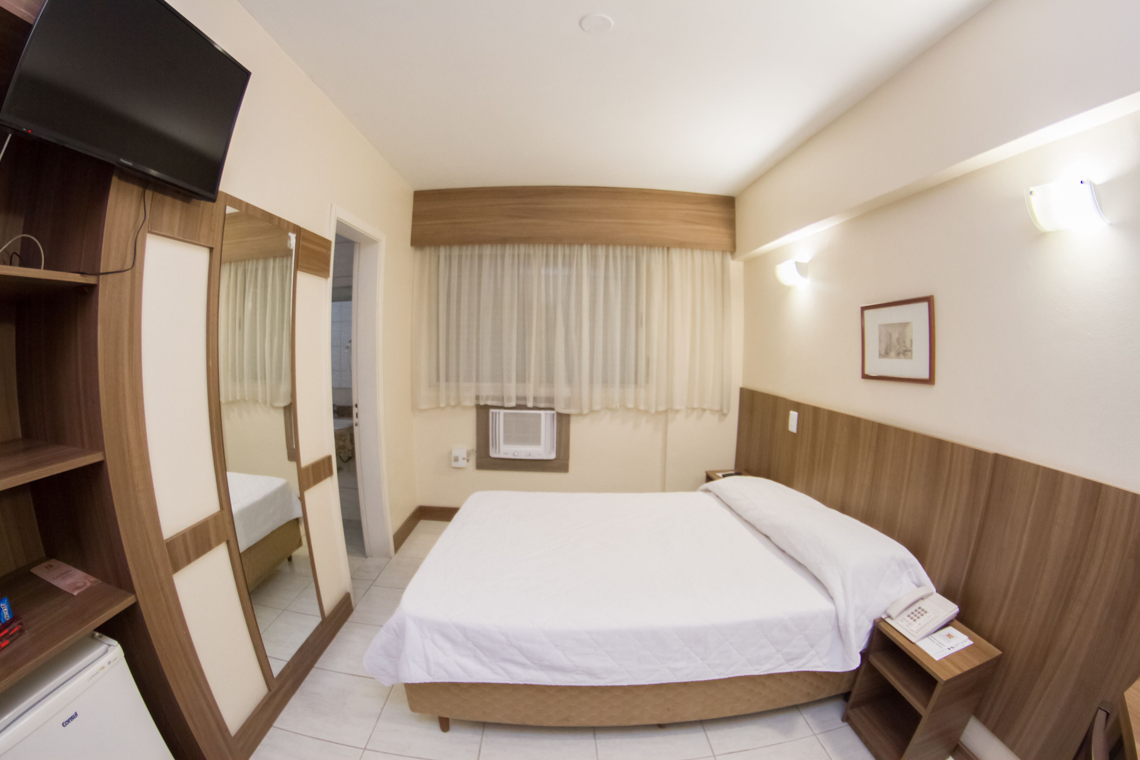 Quarto de hotel com cama, televisão, guarda-roupa, espelho, mesa de cabeceira com telefone e janela com cortinas.