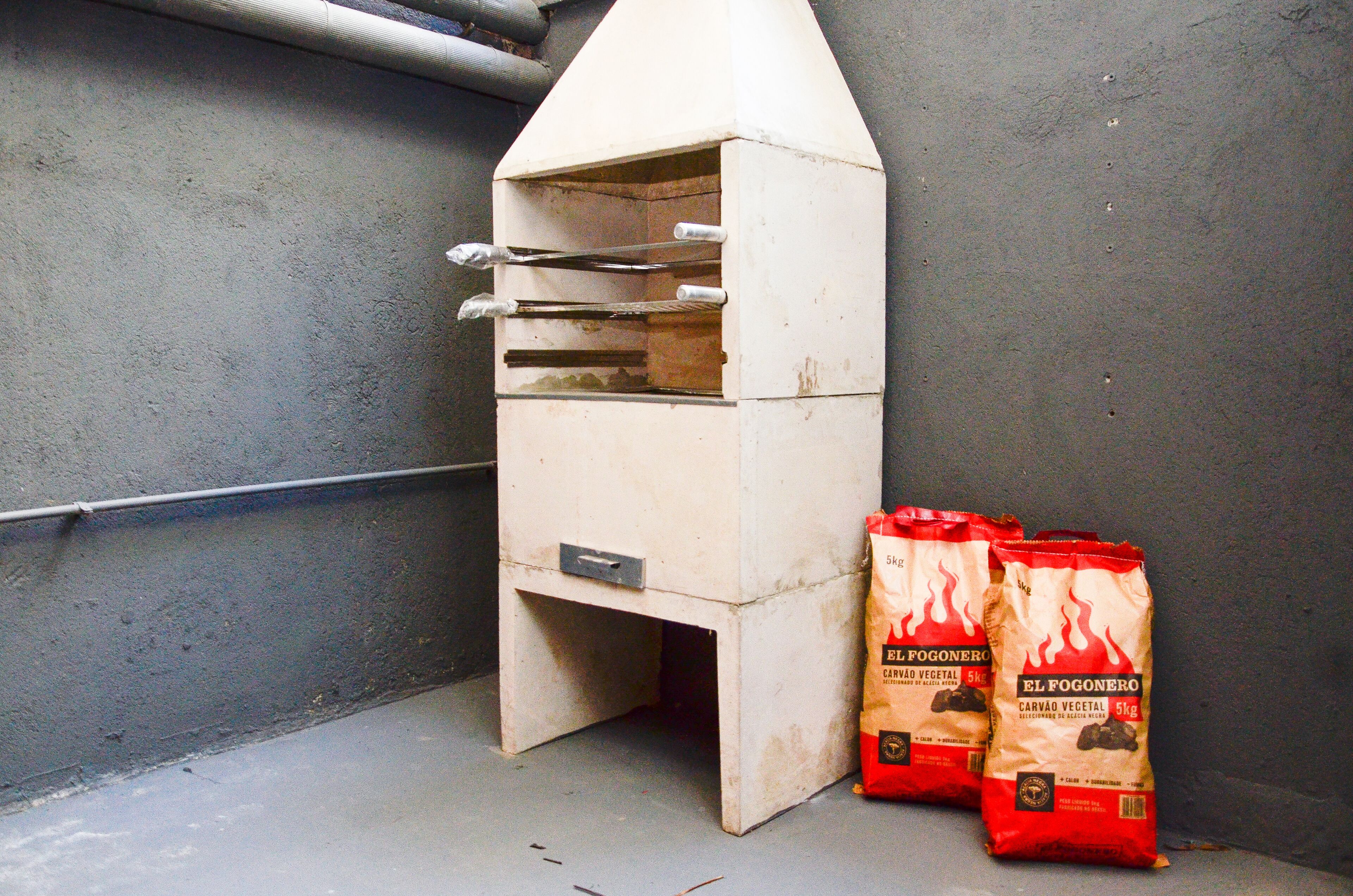 Una pequeña estructura metálica blanca con techo inclinado, posiblemente un horno portátil, junto a dos bolsas rojas con la etiqueta "El Fogonero" y un peso de carbón vegetal de 5 kg.