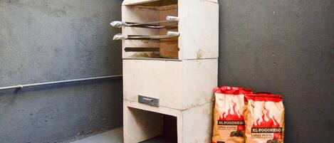Una pequeña estructura metálica blanca con techo inclinado, posiblemente un horno portátil, junto a dos bolsas rojas con la etiqueta "El Fogonero" y un peso de carbón vegetal de 5 kg.