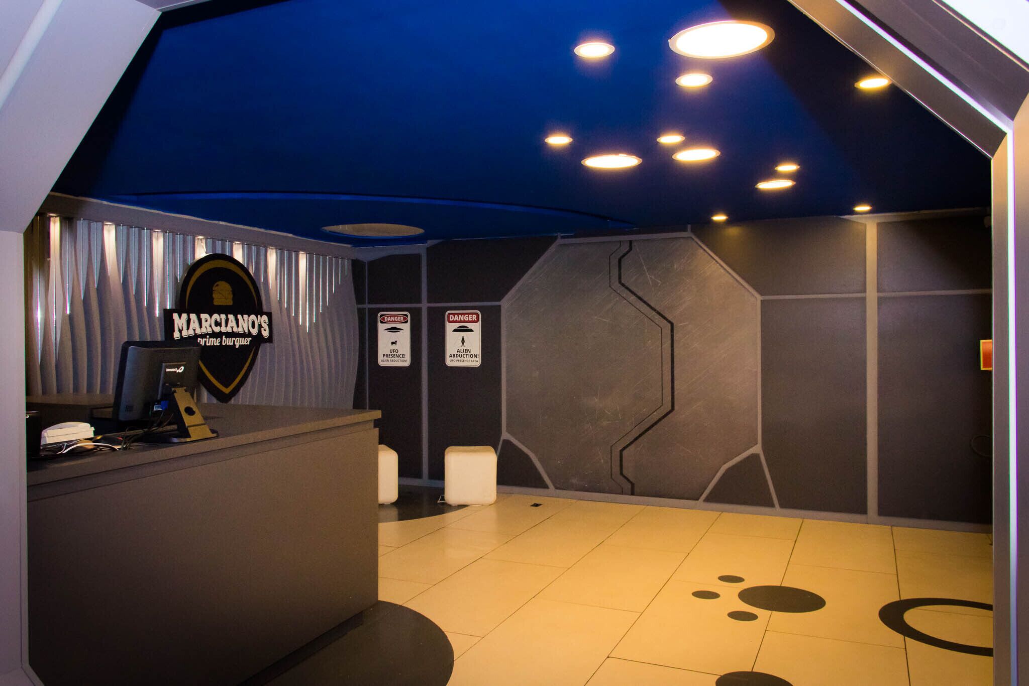 A reception area with a dark blue ceiling, a sign for 'Marciano's Prime Burger', and warning signs for 'Danger' and 'Alien Abduction'.