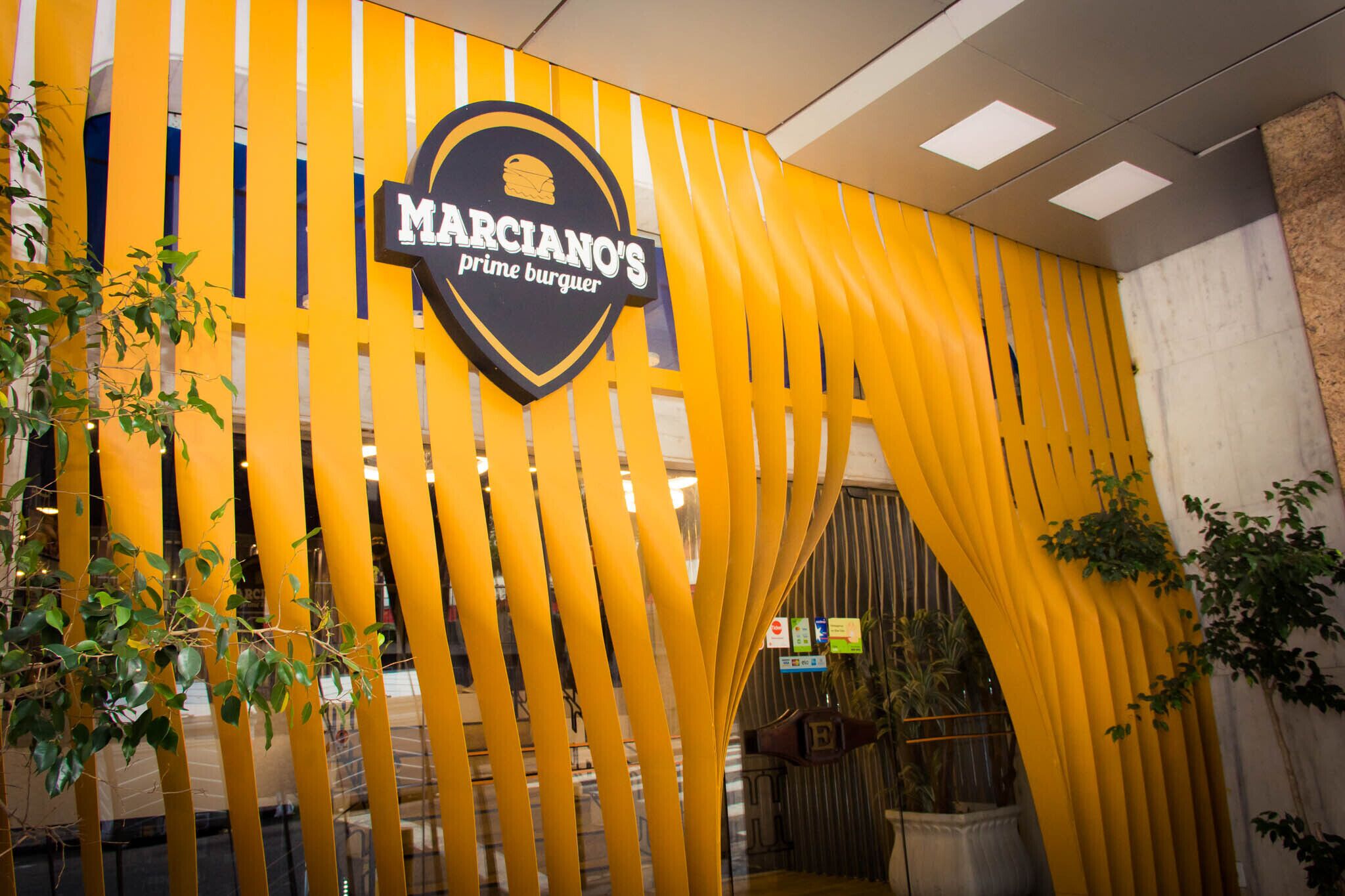 Entrada a un restaurante con fachada amarilla a rayas y un letrero que dice "Marciano's Prime Burguer".