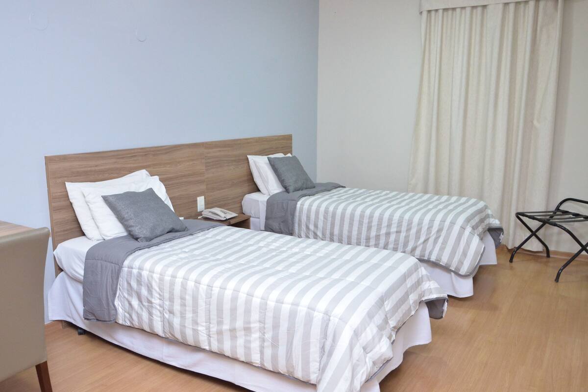A hotel room with two beds, each with striped bedding and pillows. There is a nightstand with a phone on it, and a chair is visible in the corner.