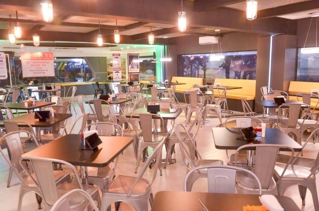 A modern restaurant with multiple tables and chairs, a green-lit counter area, and a view through large windows.
