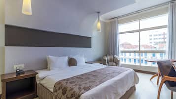 Habitació d'hotel amb un llit gran, una taula de nit amb telèfon, una cadira, una taula petita i un finestral ampli amb vista a edificis.