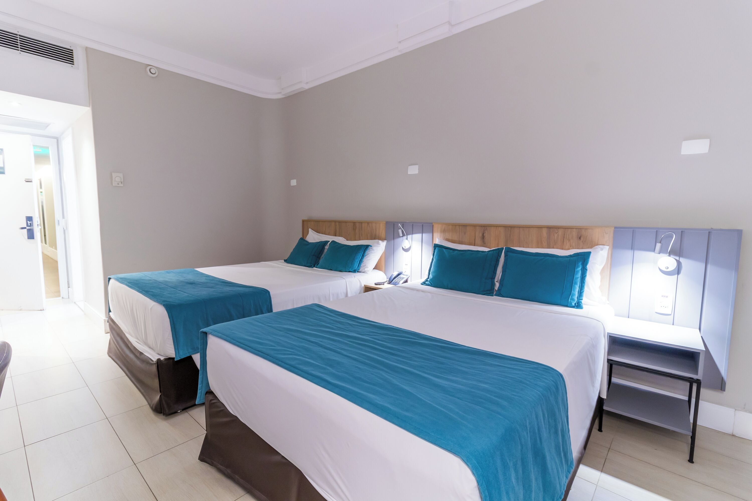 Quarto de hotel com duas camas, cada uma com cabeceira de madeira, roupa de cama branca e detalhes em tons de verde-água. Há uma mesa de cabeceira com abajur e tomada.