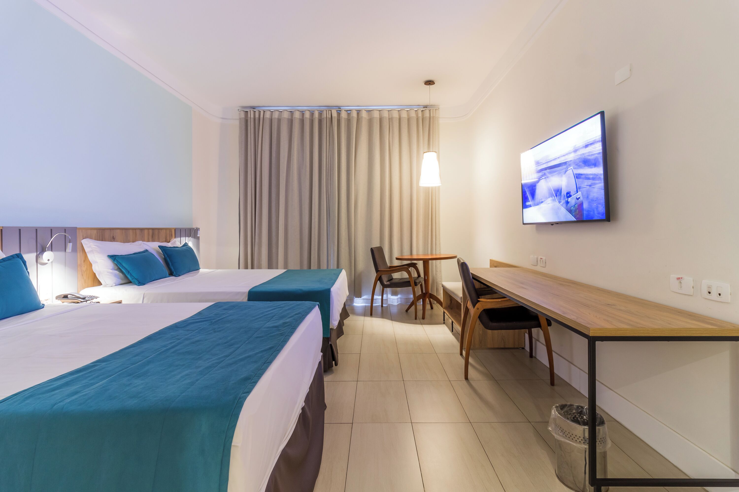 A hotel room with two beds, a desk, a chair, a television, and a window with curtains.