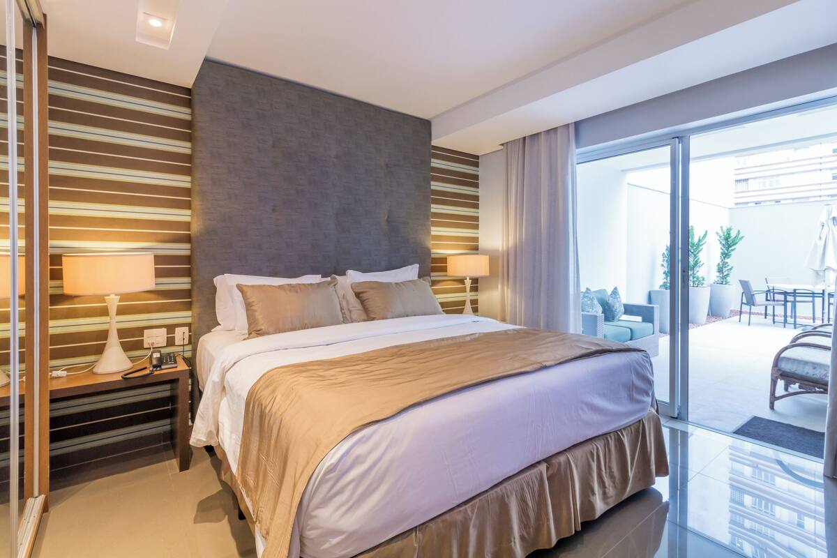 A modern hotel room with a large bed, two bedside tables with lamps, a view of the outdoor patio area through a sliding glass door.