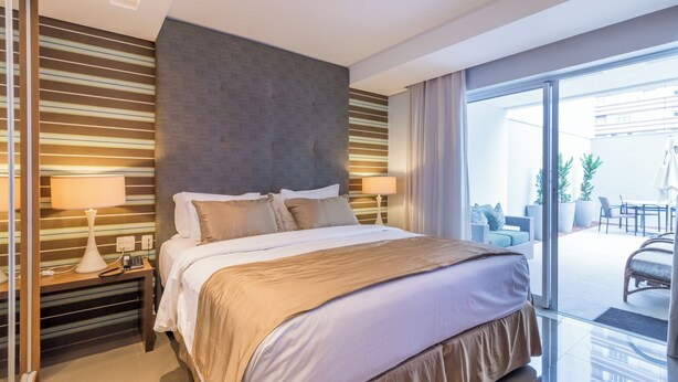 Habitación de hotel moderna con una cama grande, dos mesitas de noche con lámparas y vista al patio exterior por un ventanal corredero.