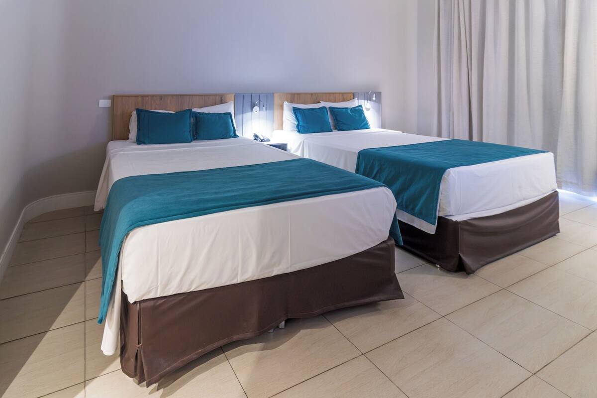 A hotel room with two beds, each with a wooden headboard, white bedding, and teal throw pillows.
