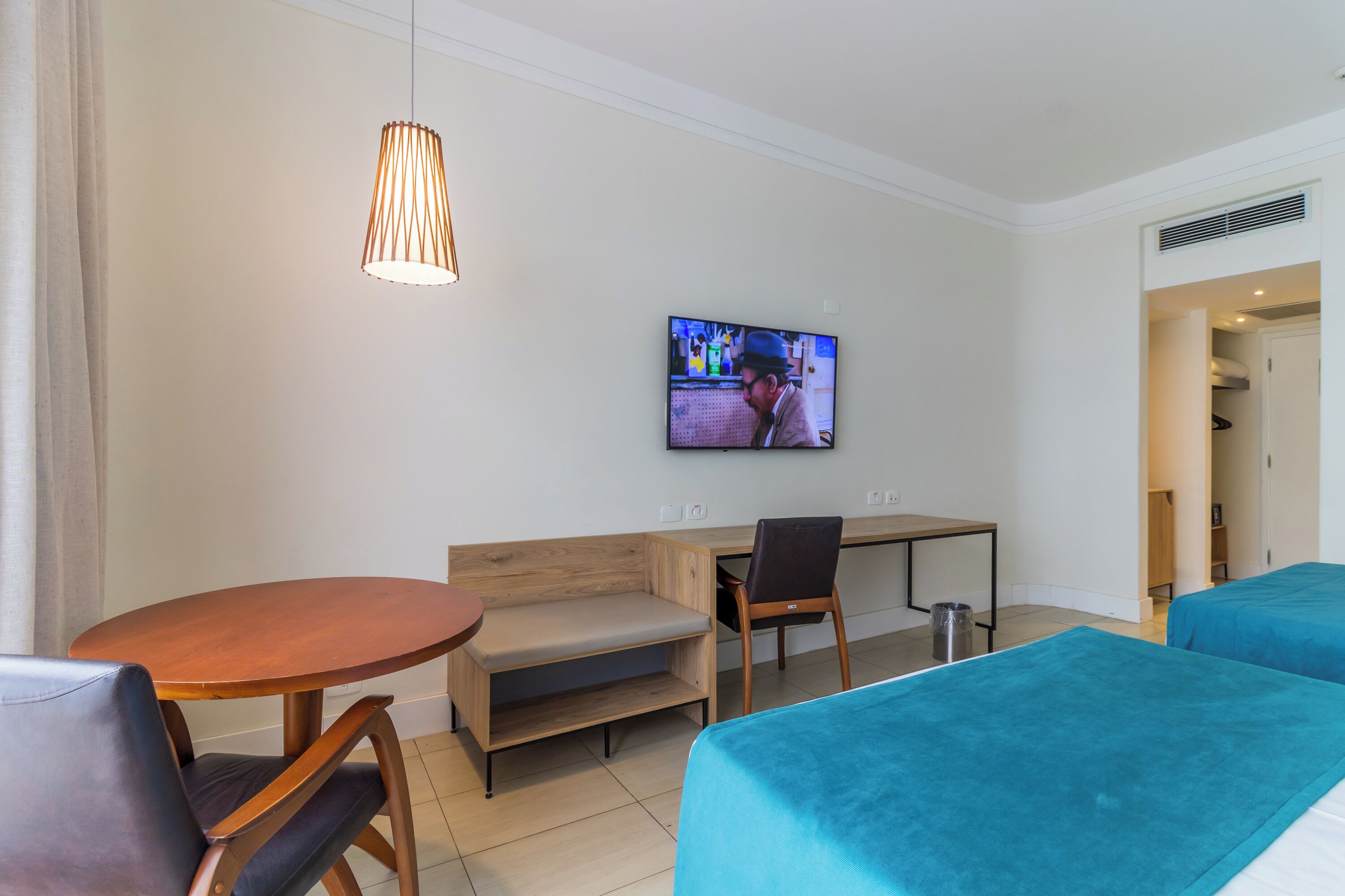 Quarto de hotel com uma escrivaninha de madeira, televisão fixada na parede, uma mesa redonda, um sofá e duas camas com roupa de cama azul.