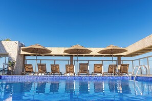 Zona de piscina con sillas de descanso y sombrillas, una pared de piedra y un cielo azul despejado.