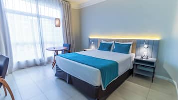 Quarto de hotel com cama grande, mesas de cabeceira, uma pequena mesa redonda e uma cadeira.