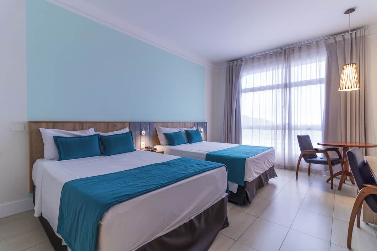 A hotel room with two beds, a wooden headboard, a table with chairs, and a large window with curtains.