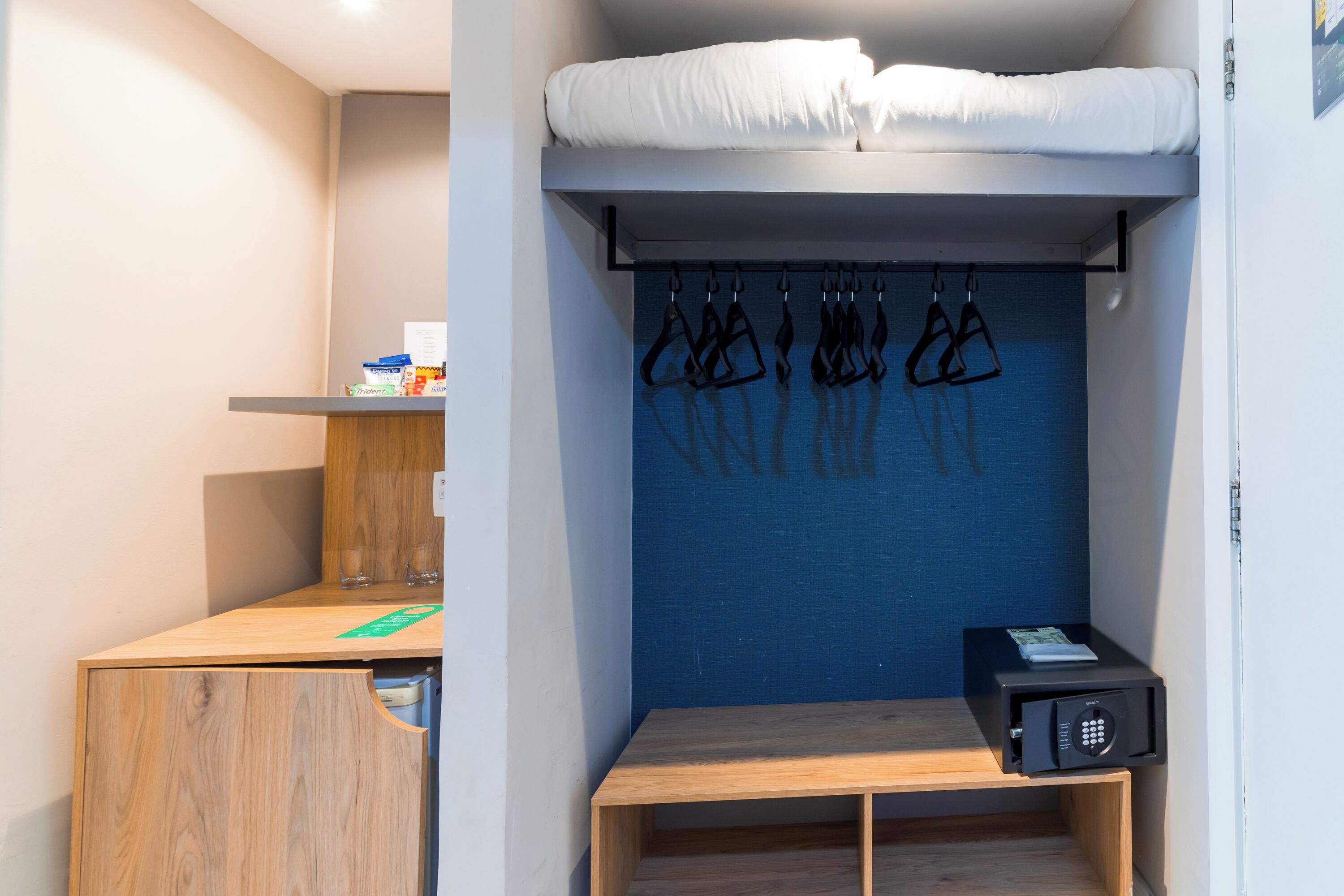 Una habitación pequeña con una cama litera, un escritorio de madera, una banca, un perchero en la pared y un dispositivo electrónico negro.