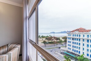 Vista desde la ventana de un hotel sobre una calle con palmeras, un edificio con ventanas azules y una playa a lo lejos.