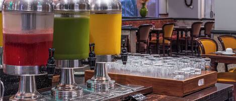 Estación de bebidas con tres dispensadores llenos de bebidas de diferentes colores, una bandeja de vasos y una máquina de café.