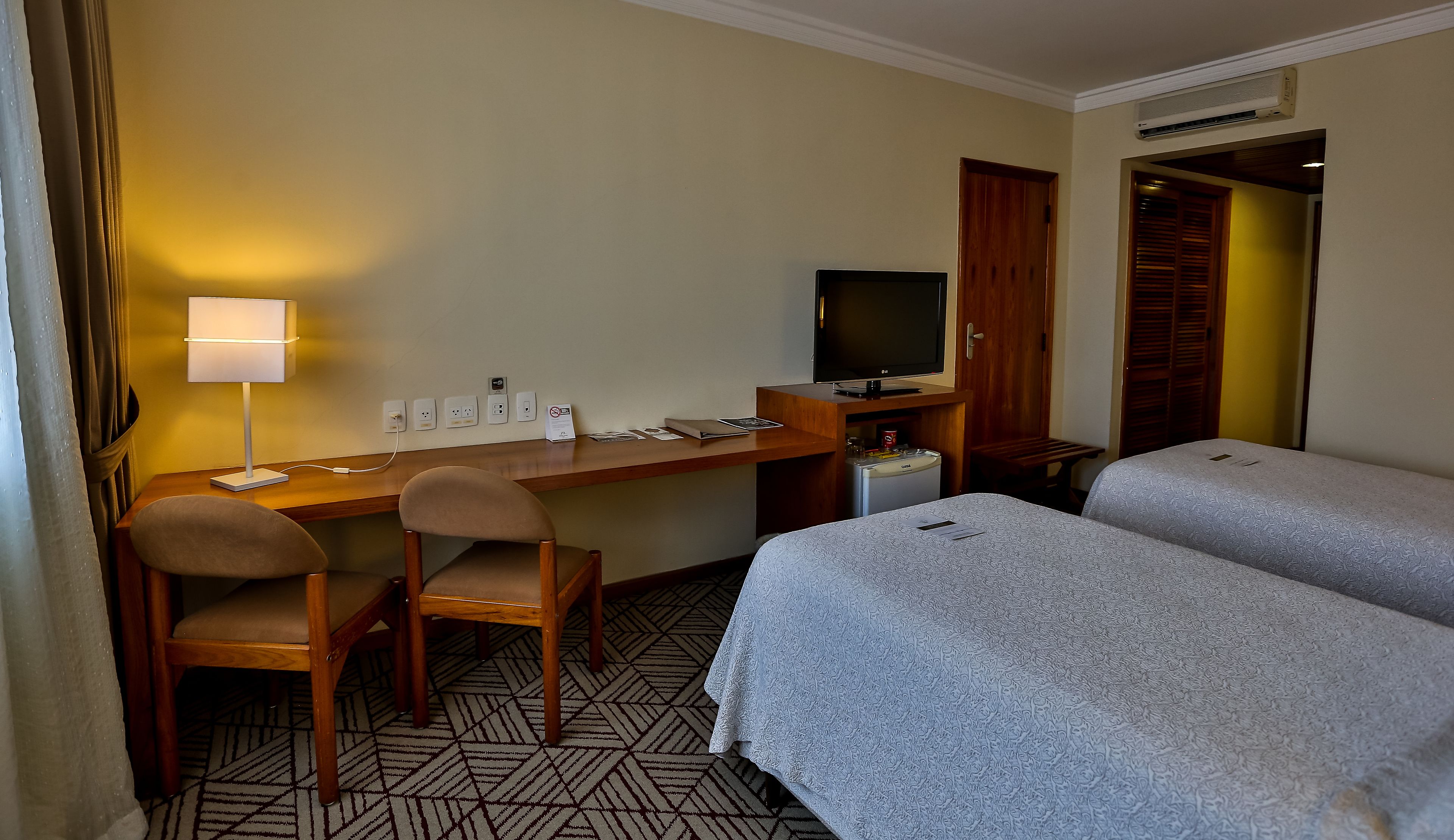 A hotel room with a desk, two chairs, a television, a minibar, and two beds.