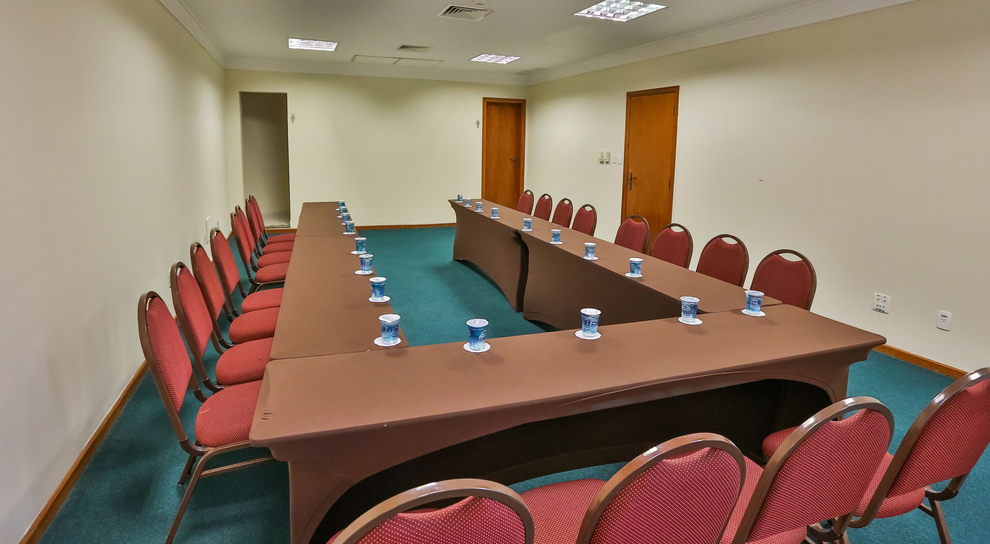 Sala de conferência com uma mesa comprida, cadeiras vermelhas e garrafas de água azuis.