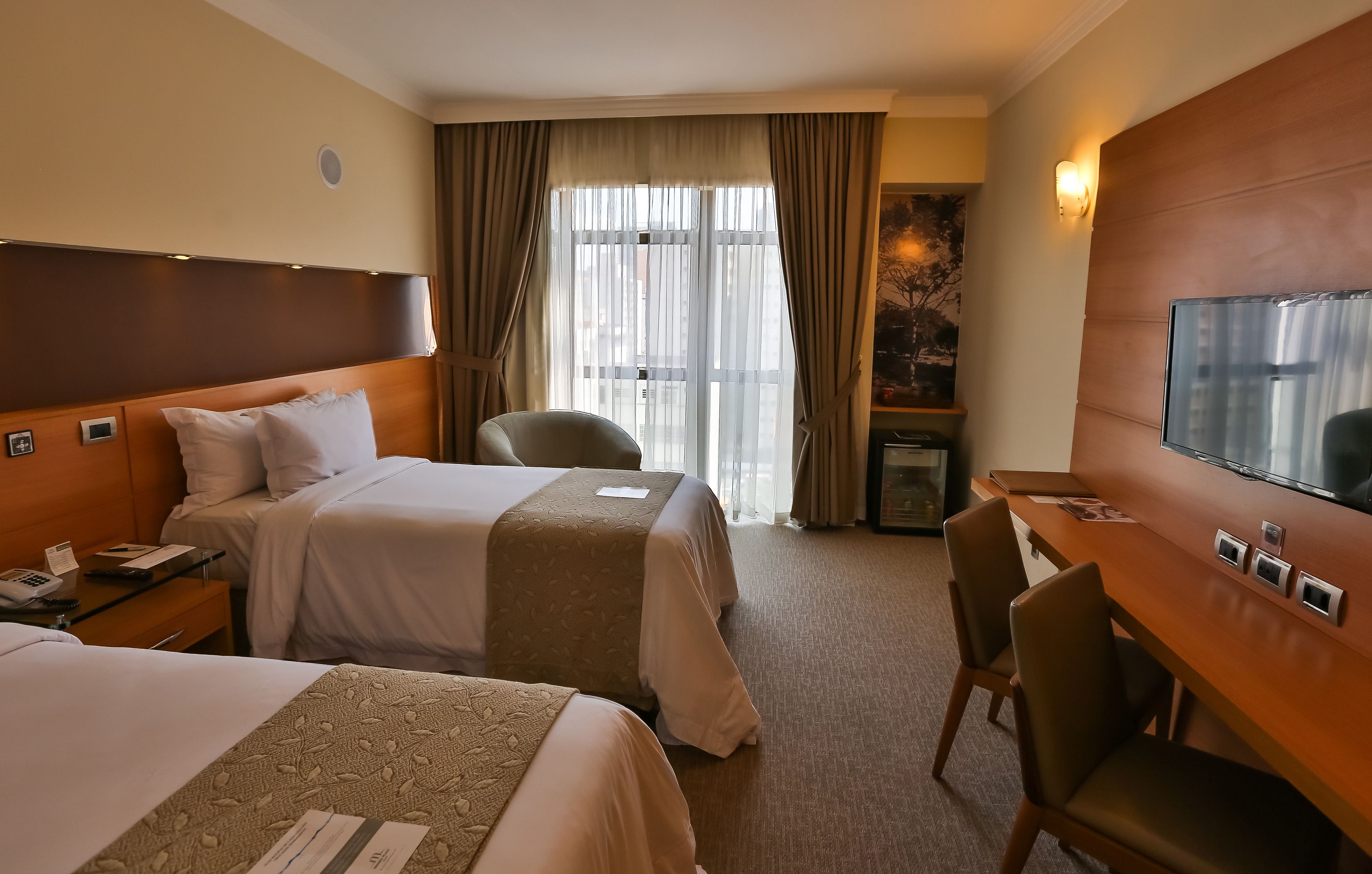 A hotel room with two beds, a desk, a chair, a television, and a window with curtains.