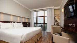 Habitación de hotel con una cama grande, televisión, una silla y vista a un paisaje urbano a través de un ventanal.