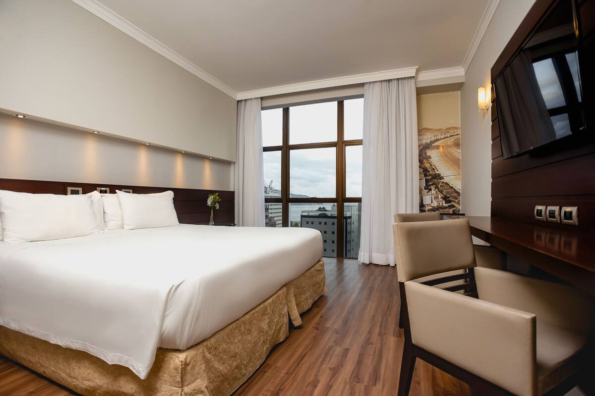 Ein Hotelzimmer mit einem großen Bett, einem Fernseher, einem Sessel und Blick auf eine Stadtlandschaft durch ein großes Fenster.
