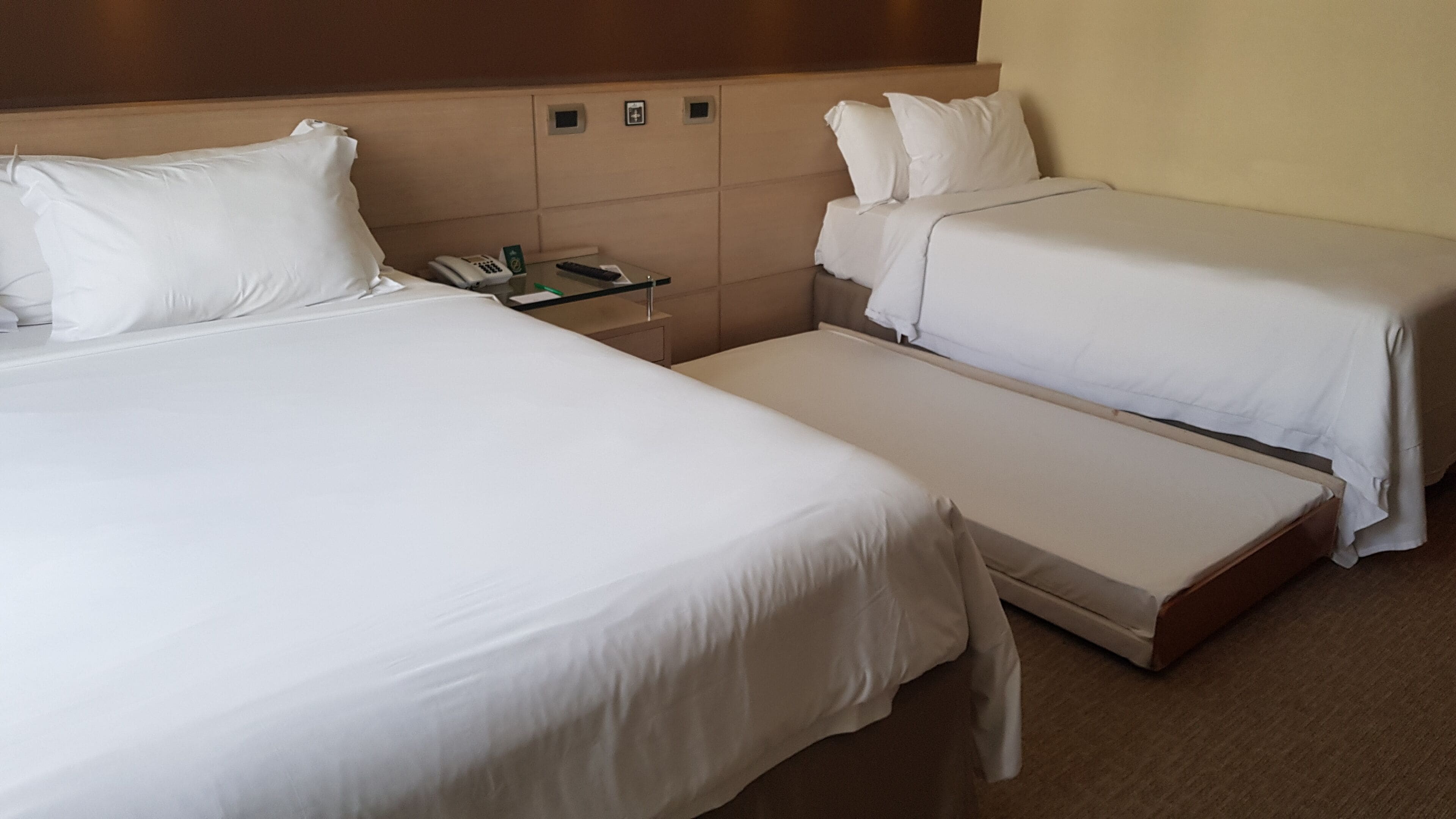 Hotellihuone, jossa on kaksi erillistä sänkyä, yöpöytä, jossa on puhelin, ja pieni työpöytä, jossa on lamppu.