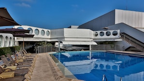 Una piscina con sillas de descanso y sombrillas, rodeada por un edificio moderno con ventanales circulares y una escalera.
