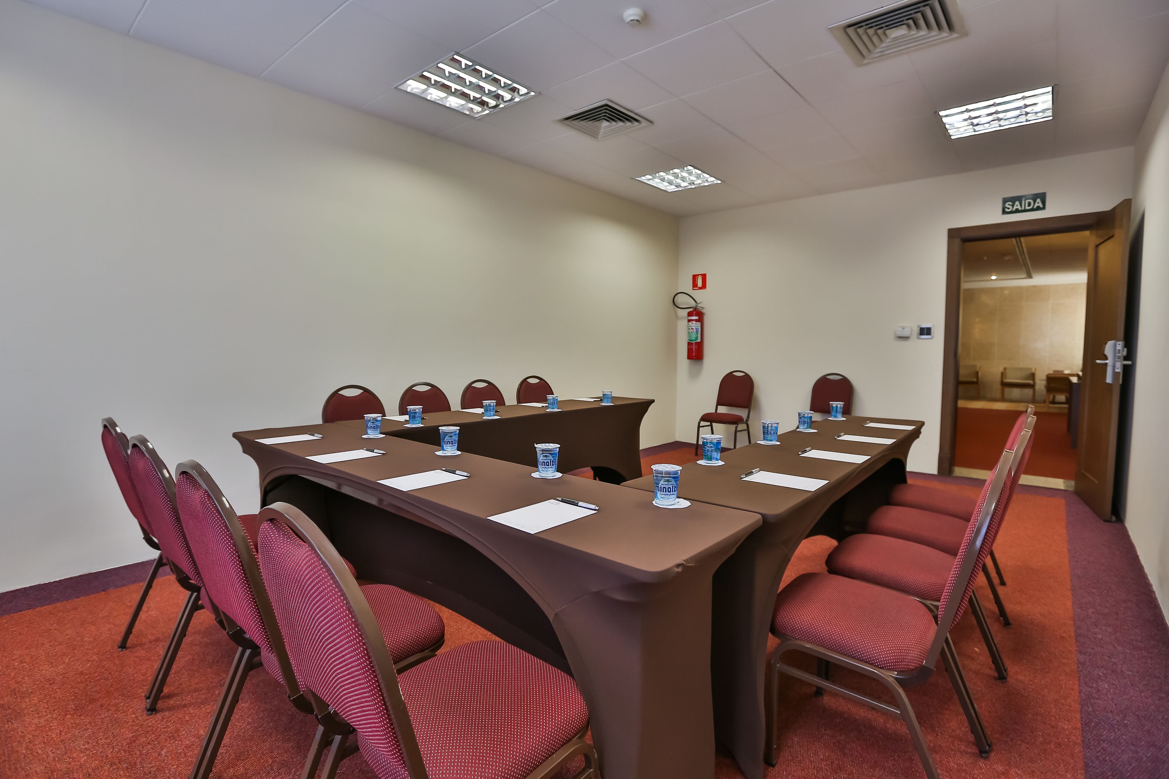 A conference room with a long table, red chairs, and a fire extinguisher on the wall.