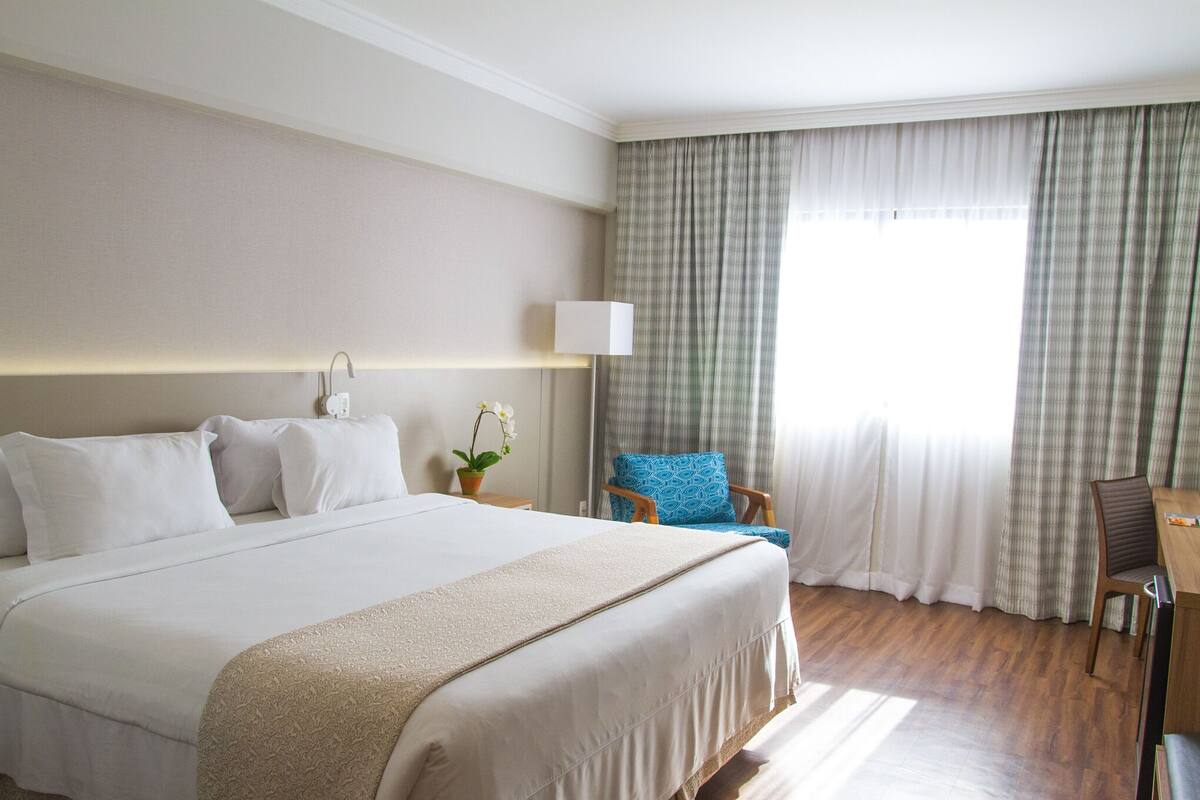 Ein Hotelzimmer mit einem großen Bett, einem Schreibtisch, einem Sessel, einer Lampe und einem fenster mit Vorhängen.
