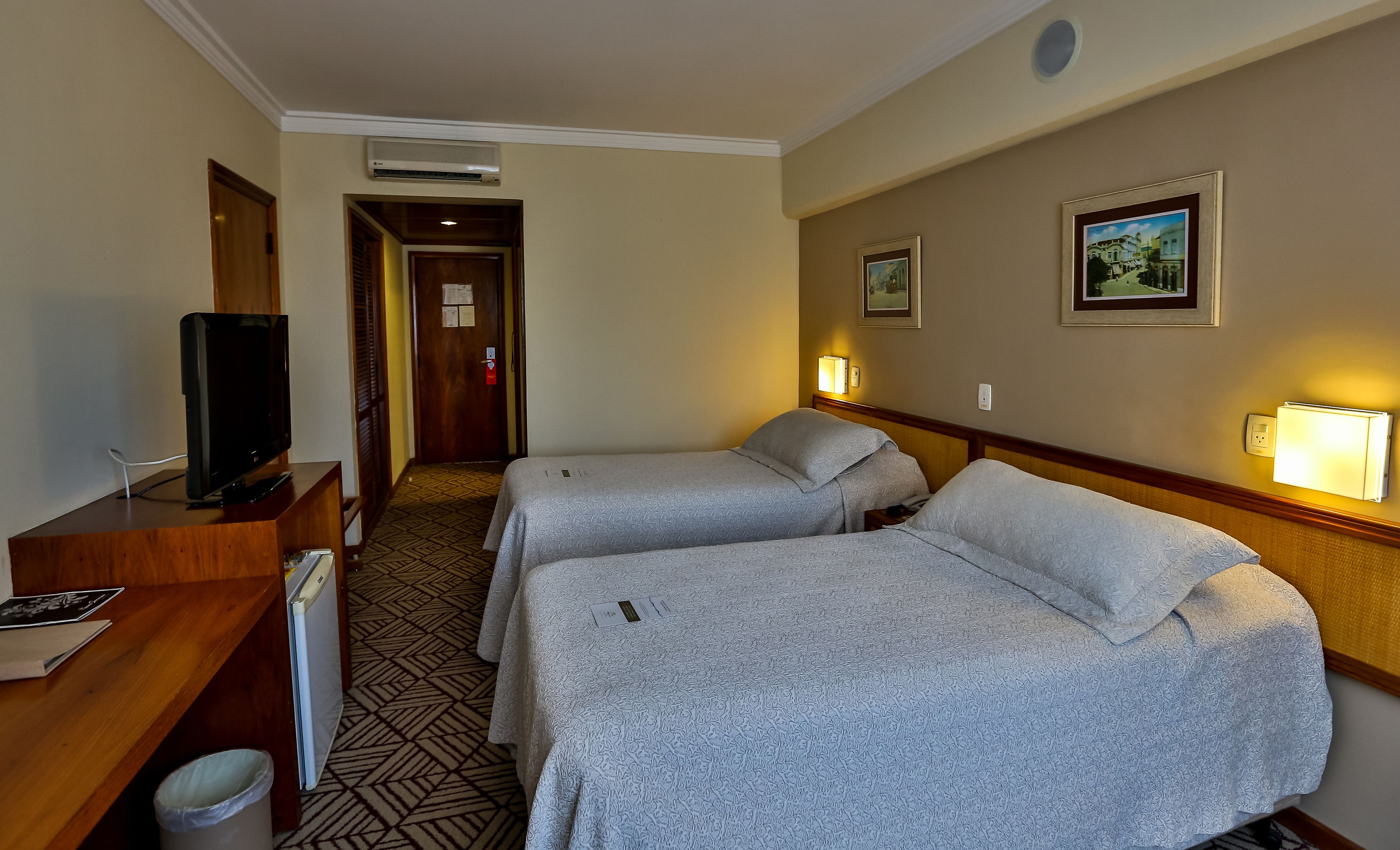 Une chambre d’hôtel avec deux lits, un bureau avec une télévision et un couloir menant à une porte.