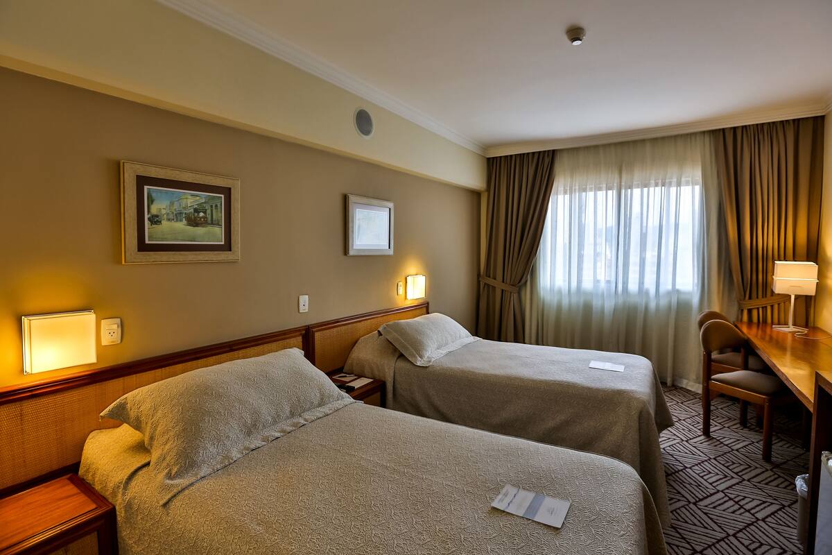 Ein Hotelzimmer mit zwei Betten, einem Schreibtisch, einem Stuhl, einer Lampe, einem Gemälde und einem fenster mit Vorhängen.
