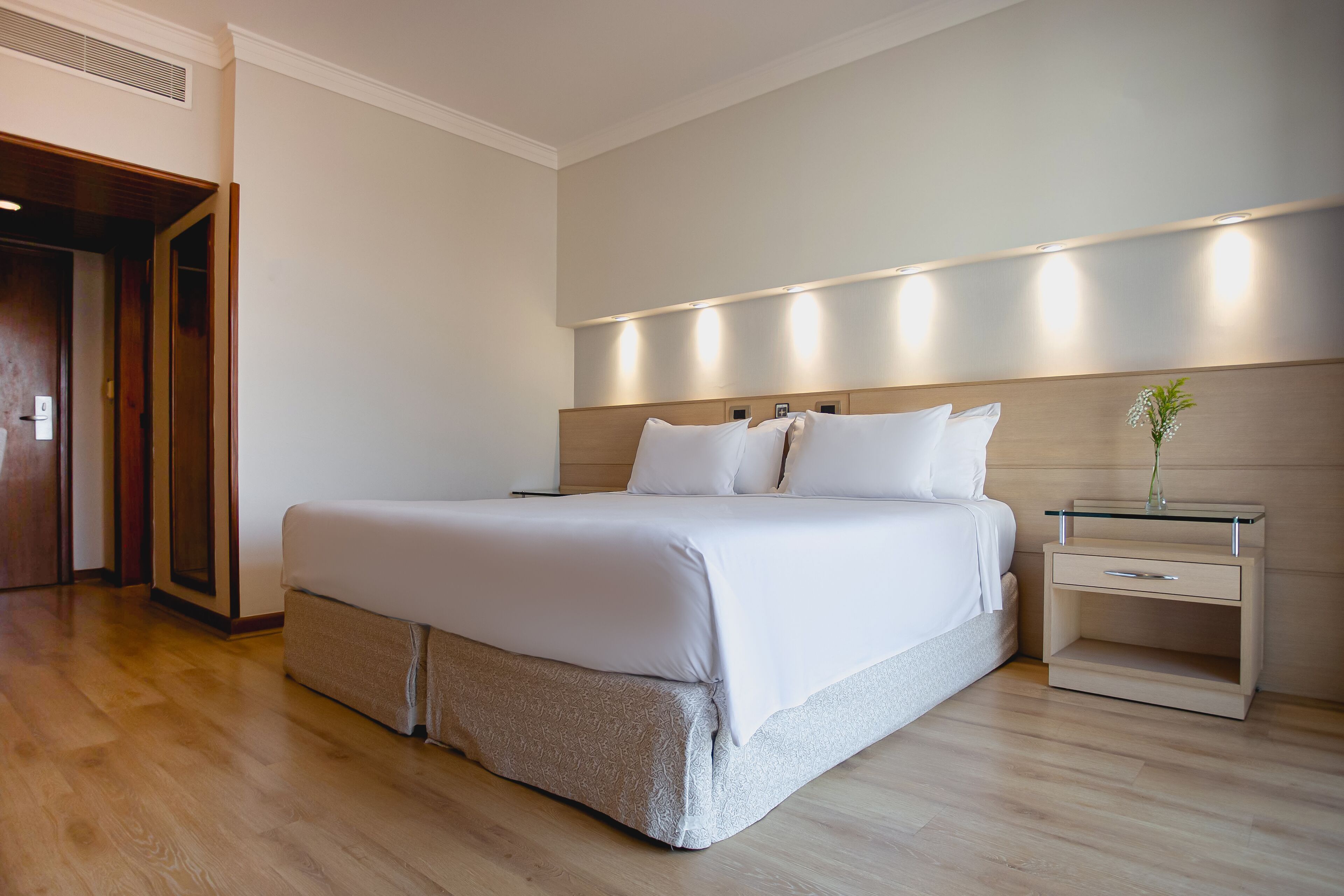 Quarto de hotel moderno com uma cama grande, uma mesa de cabeceira com um vaso e piso de madeira.