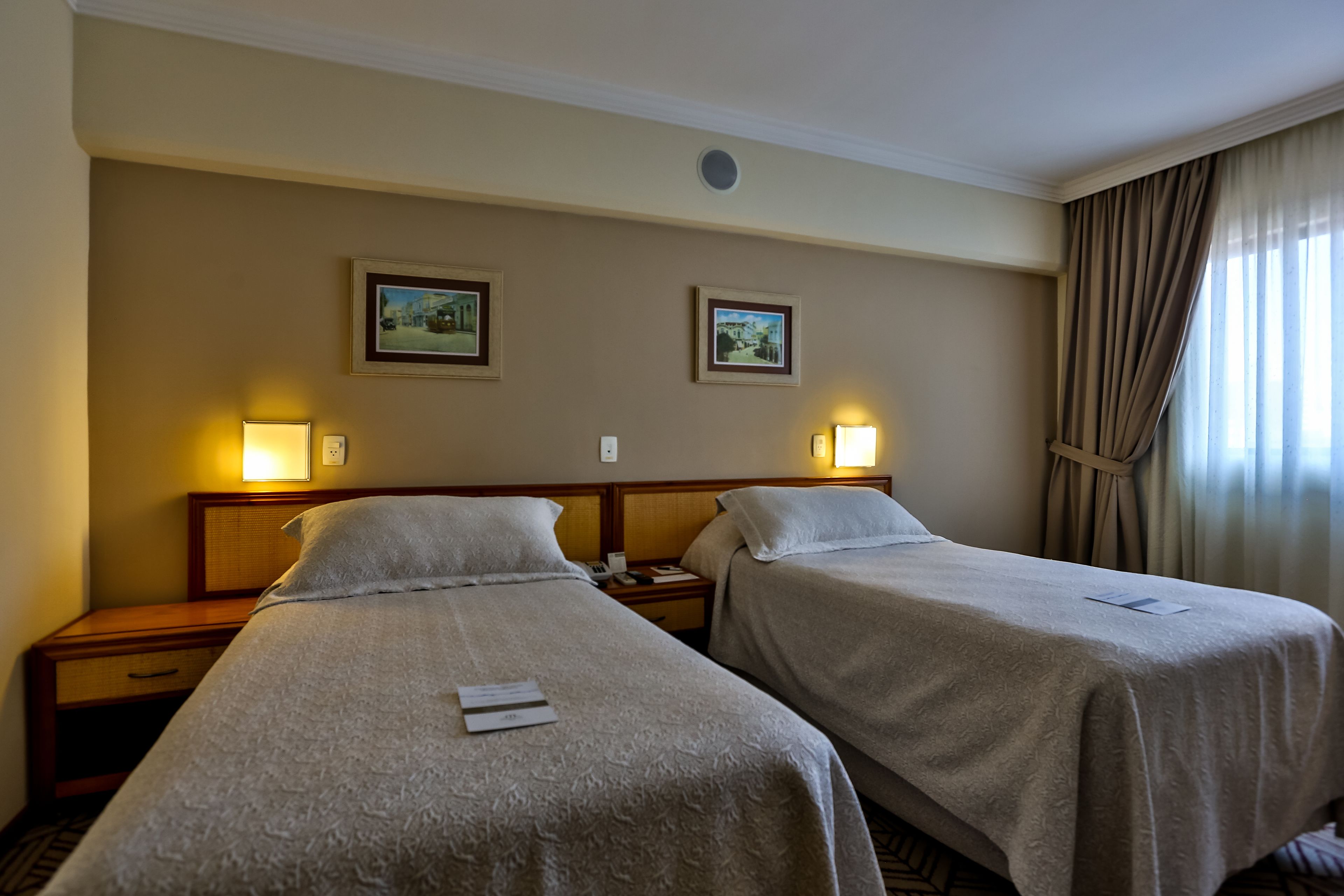 Quarto de hotel com duas camas de solteiro, uma mesa de cabeceira com abajur e duas fotografias emolduradas na parede.