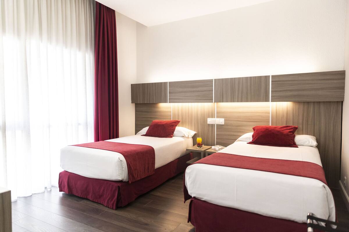 A hotel room with two beds, each with red bedding and pillows, a wooden headboard, and a nightstand with a lamp and a phone.