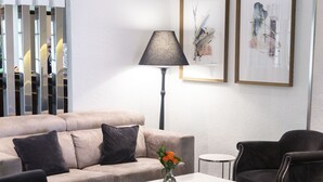 A modern living room with a beige sofa, two black armchairs, a white coffee table, and a floor lamp. There are two framed artworks on the wall and a decorative mirror on the left.