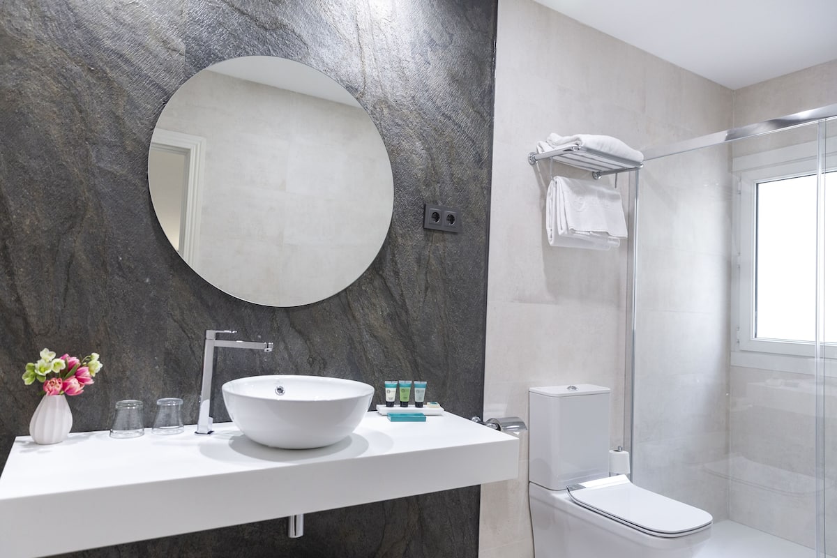A modern bathroom with a large round mirror, a white sink, and a glass-enclosed shower.