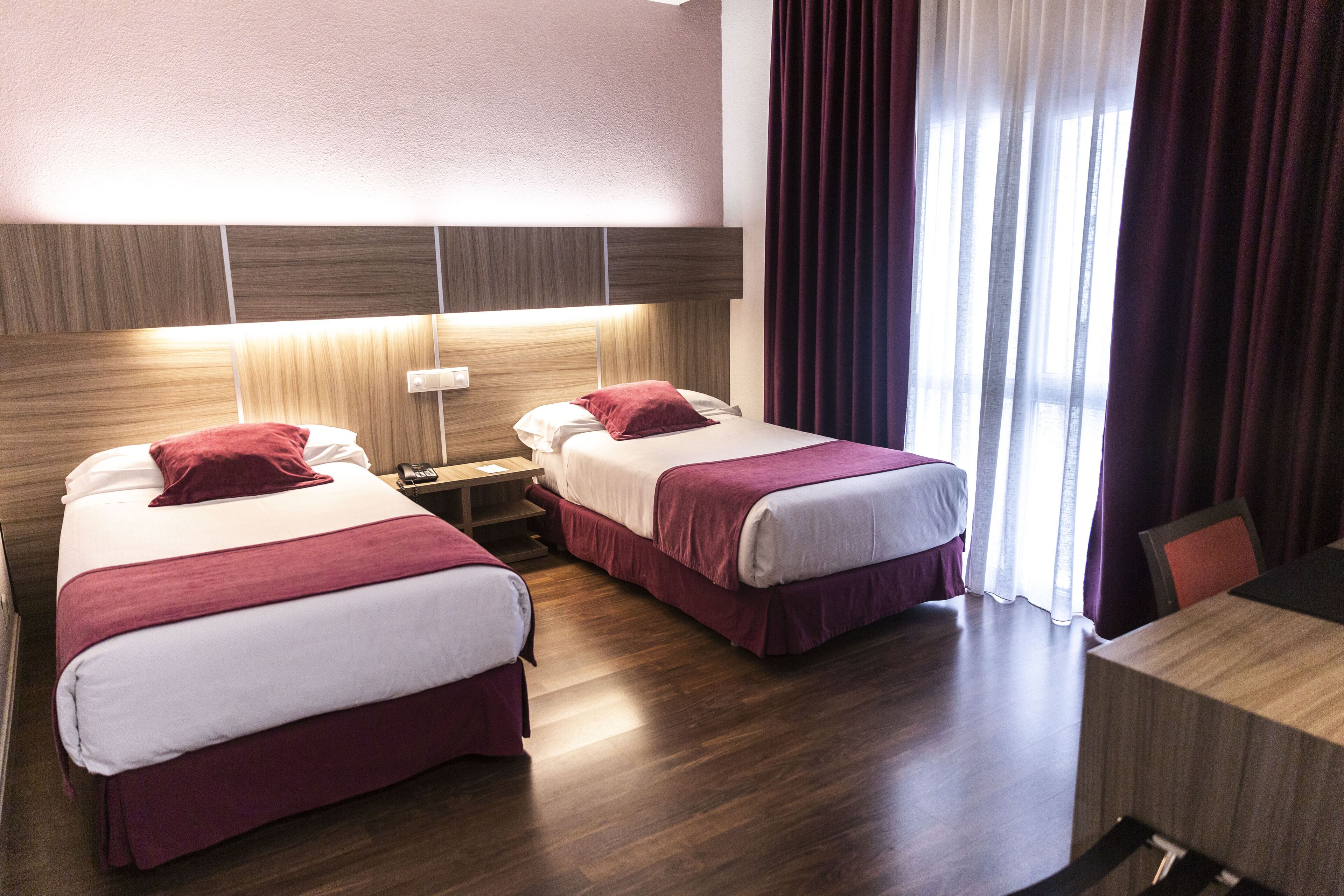 A hotel room with two beds, a bedside table with a lamp, a desk, and a window with curtains.