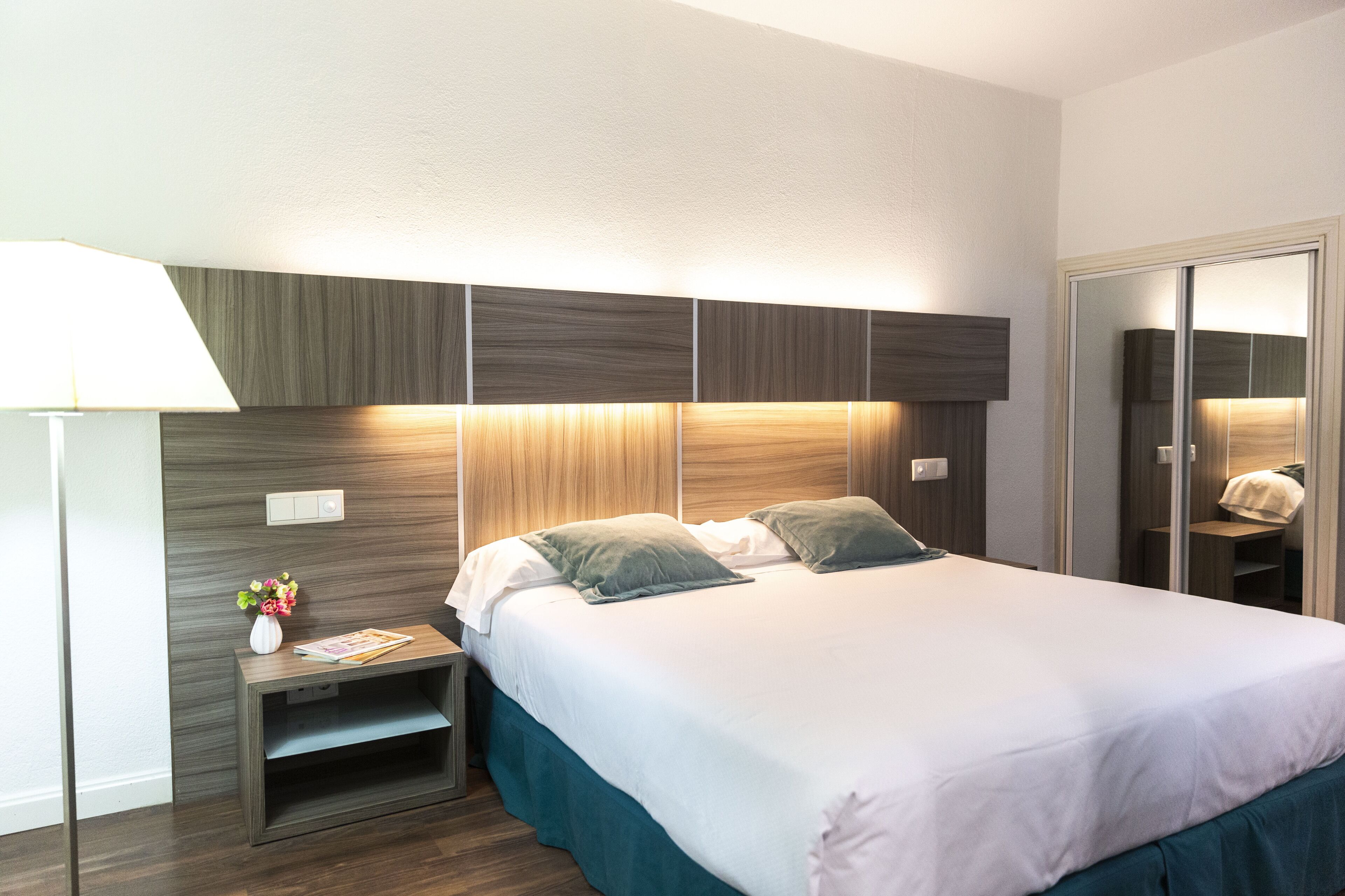 A hotel room with a bed, bedside table, lamp, and a mirror.