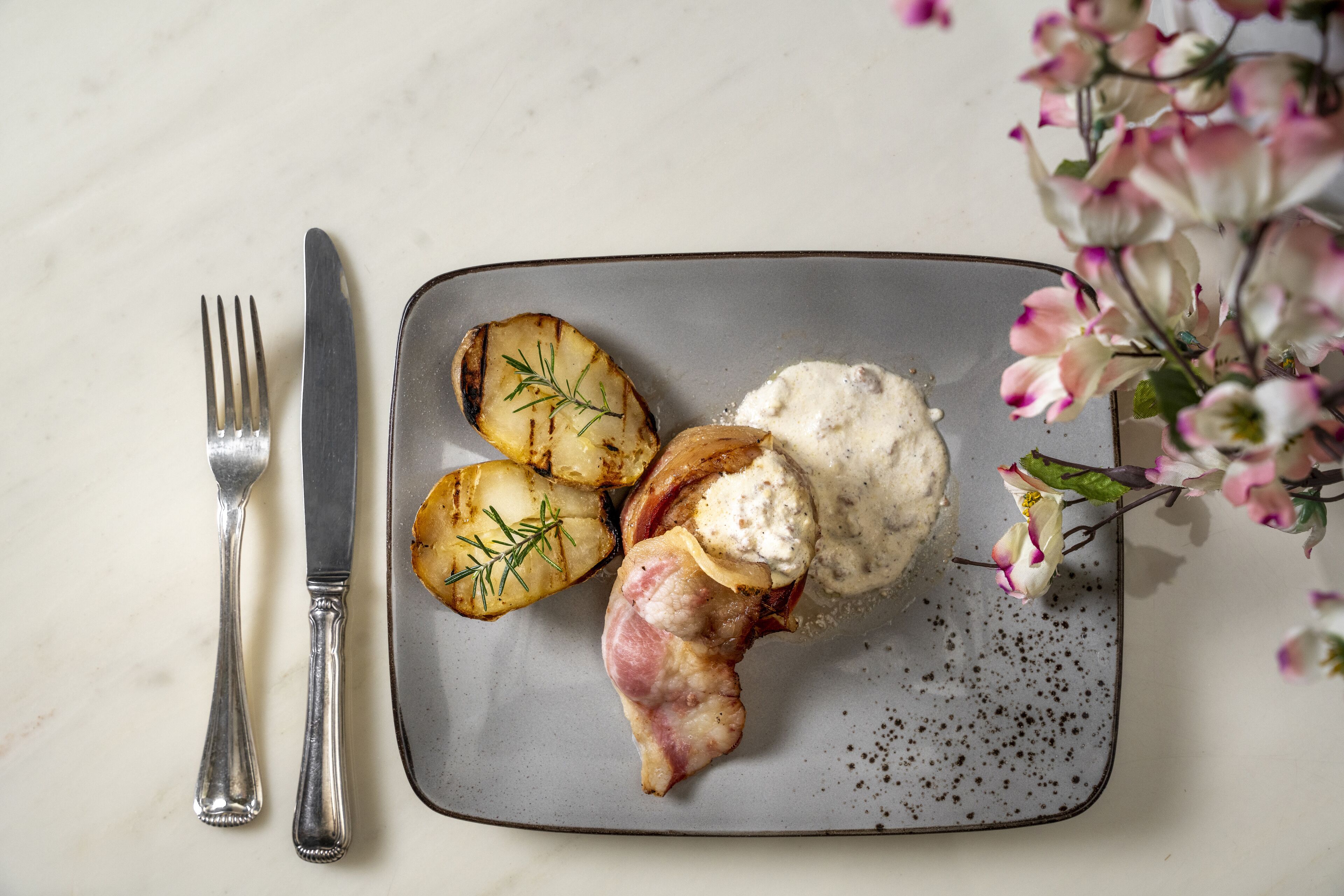 A plate with roasted potatoes, a piece of bacon, and a creamy sauce, accompanied by a fork and knife on a marble surface.