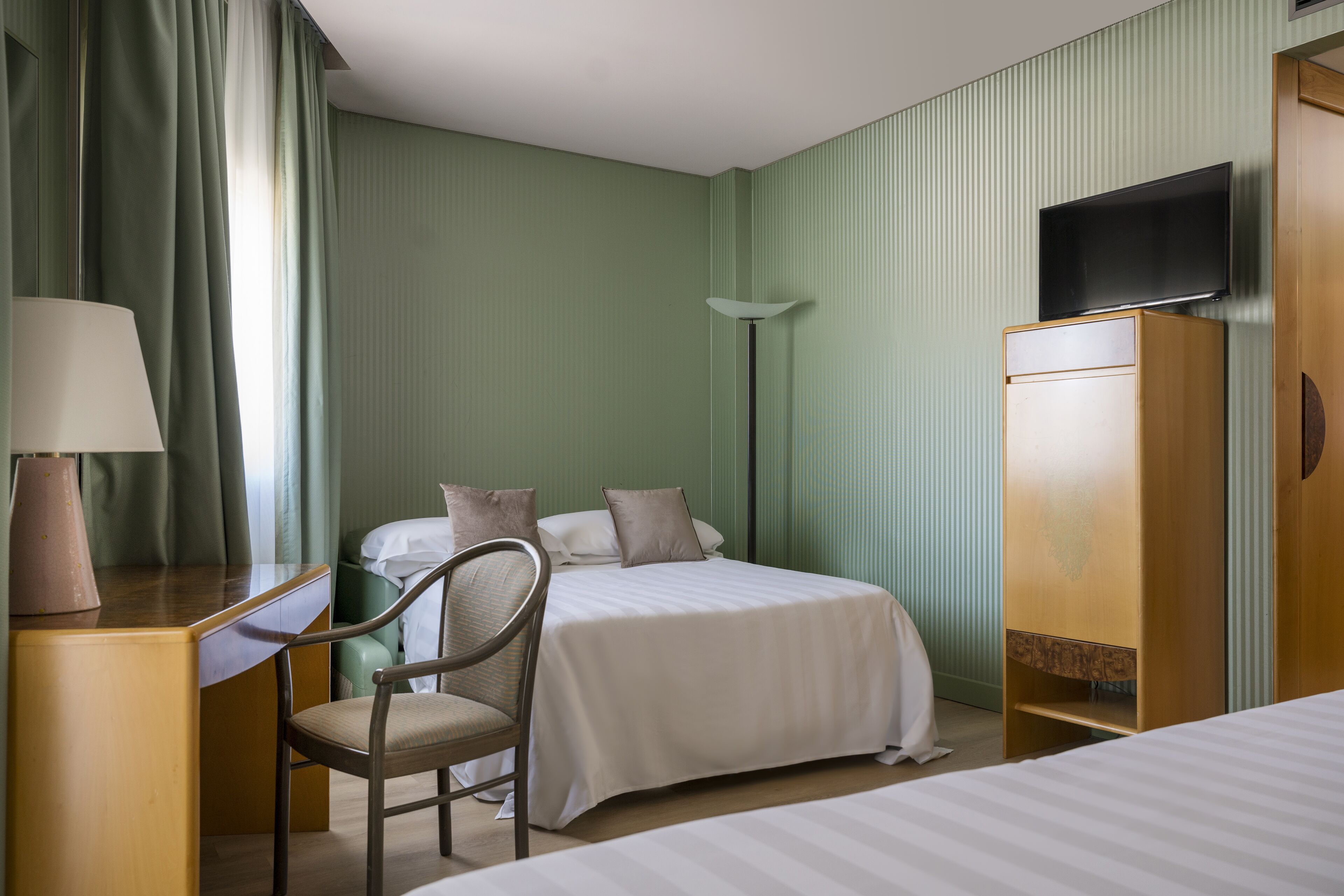 A hotel room with a bed, a desk with a lamp, a chair, a wardrobe, and a television on the wall.