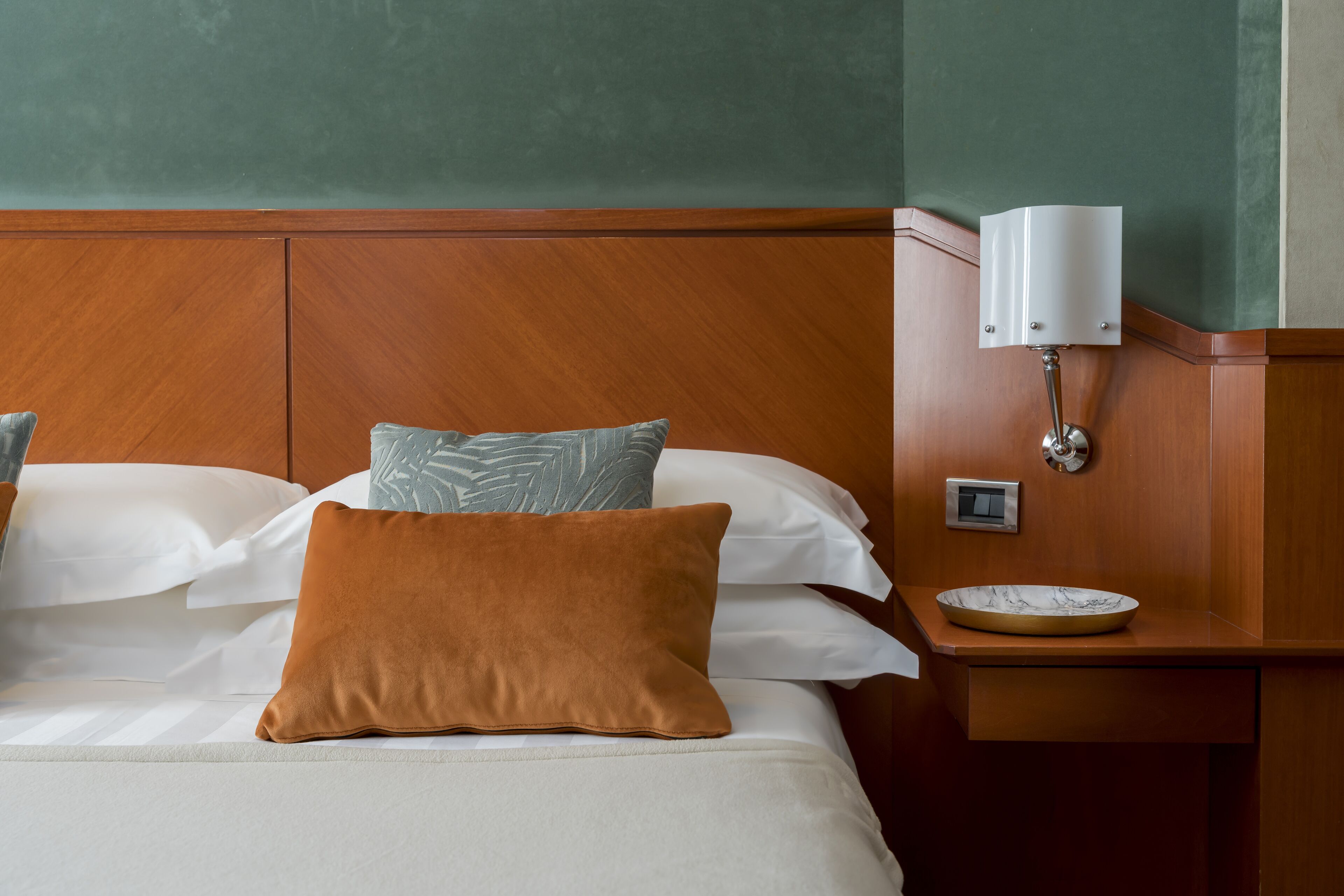 Ein ordentlich bezogenes Bett mit weißen und braunen Kissen, ein hölzernes Kopfteil und ein Nachttisch mit Lampe und Schale.