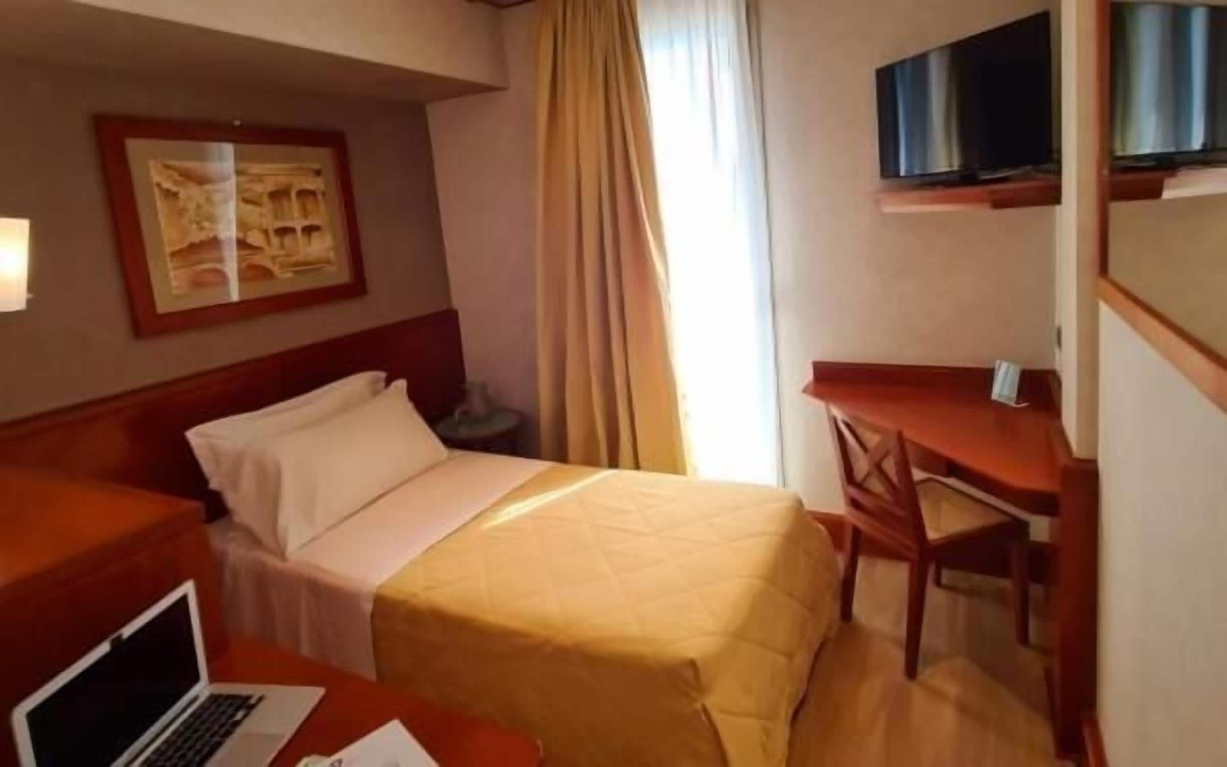 Una habitación de hotel con cama, escritorio, silla, televisor y ordenador portátil.