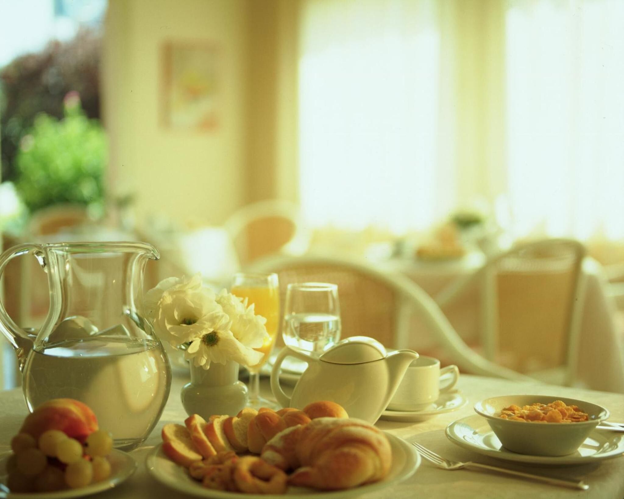 A breakfast setting with a pitcher, teapot, cups, a bowl of cereal, and pastries on a table.