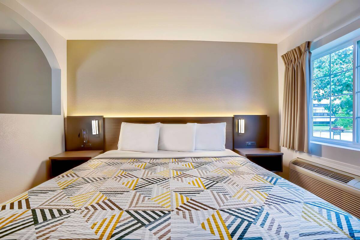 A hotel room with a large bed, two bedside tables, a headboard with integrated lighting, a window with curtains, and a view of the outdoors.