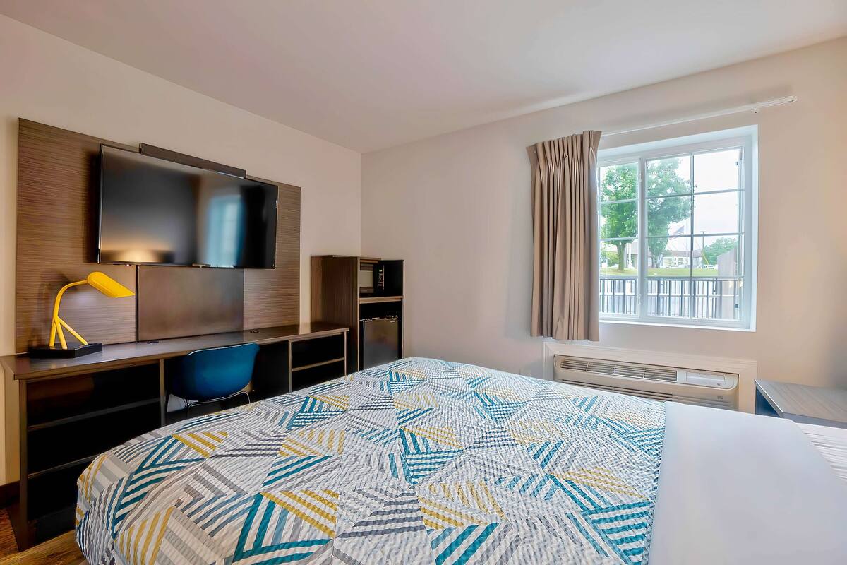 A hotel room with a bed, a desk with a computer, a TV, and a window with curtains.