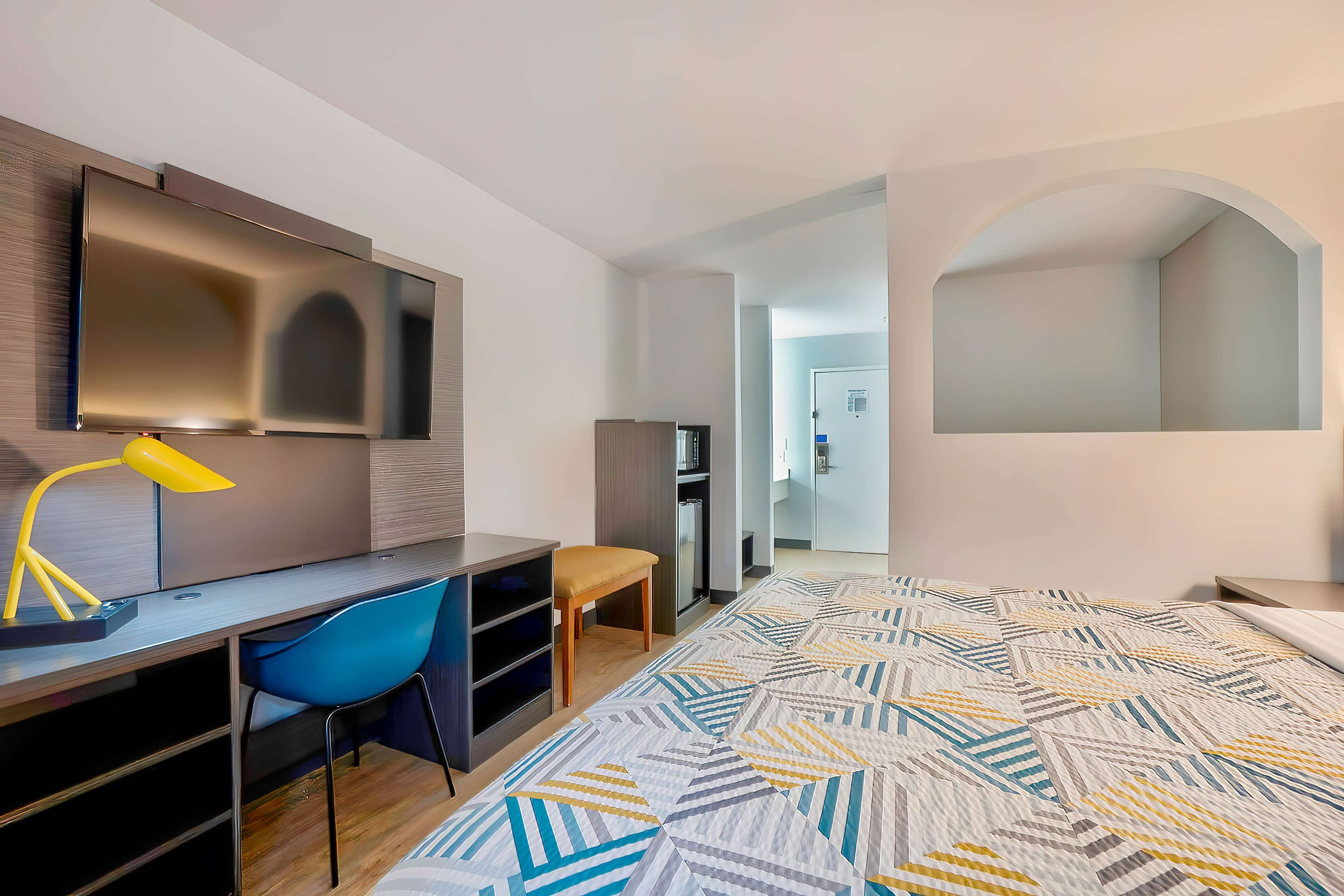 Una habitación de hotel moderna con una cama grande, un escritorio con silla, un televisor en la pared y un armario.