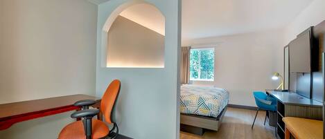 A hotel room with a desk, chair, bed, and TV.