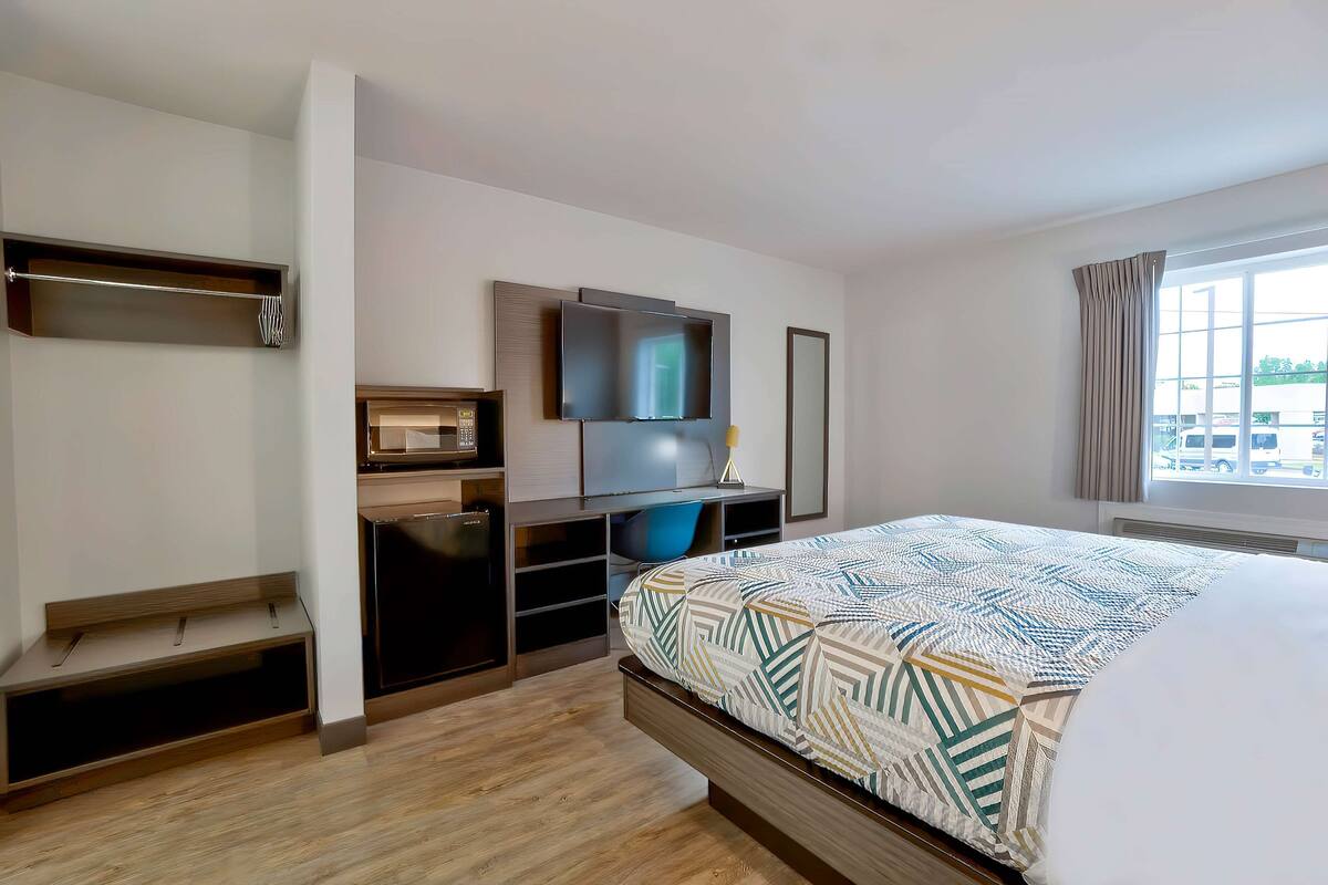 A modern hotel room with a bed, a desk with a flat-screen TV, a built-in bench, and a large window.