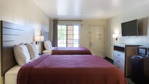 Quarto de hotel com duas camas, uma televisão, uma escrivaninha com computador e uma porta.